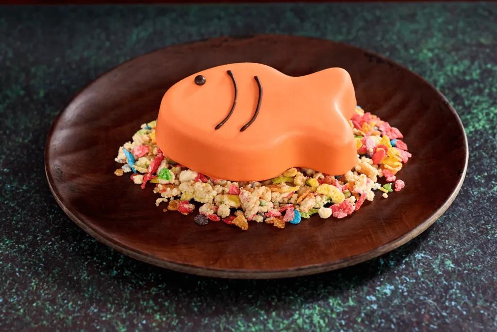 A chocolate mousse fish which is bright orange with brown details on top of rice Krispy cereal bites.