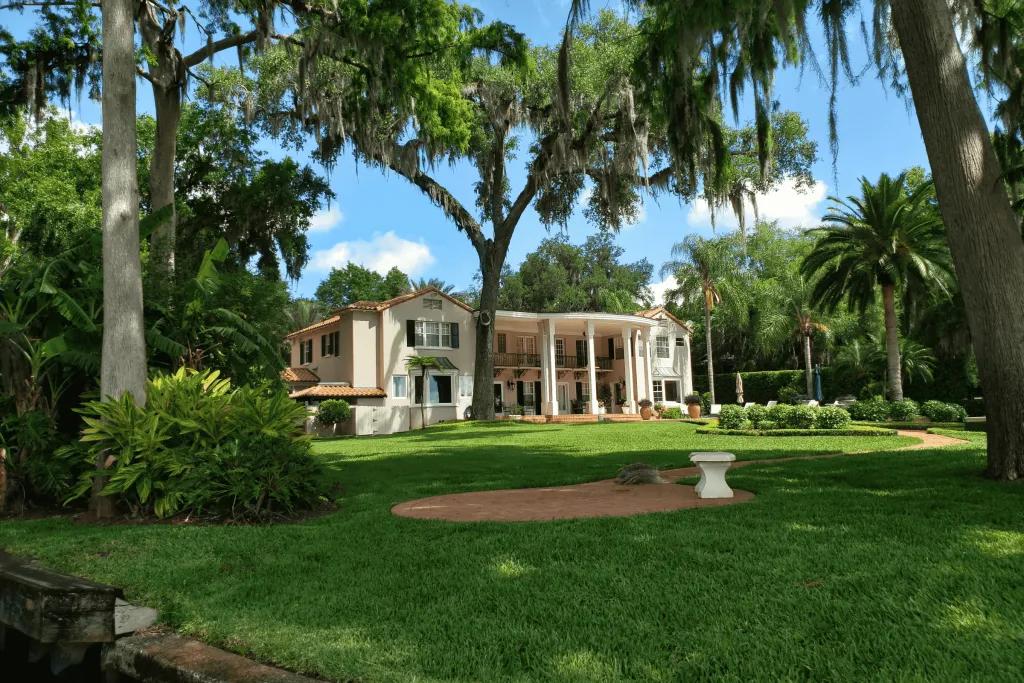 A large cream mansion with a lot of garden space, with trees and small white benches.