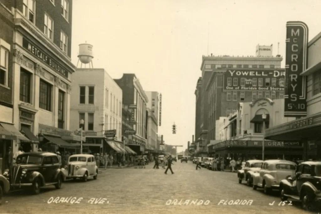 A black and white image of a street with pedestrians crossing in front of old fashioned beetle cars. There is old fashioned buildings, one with a water tower on top of it. 