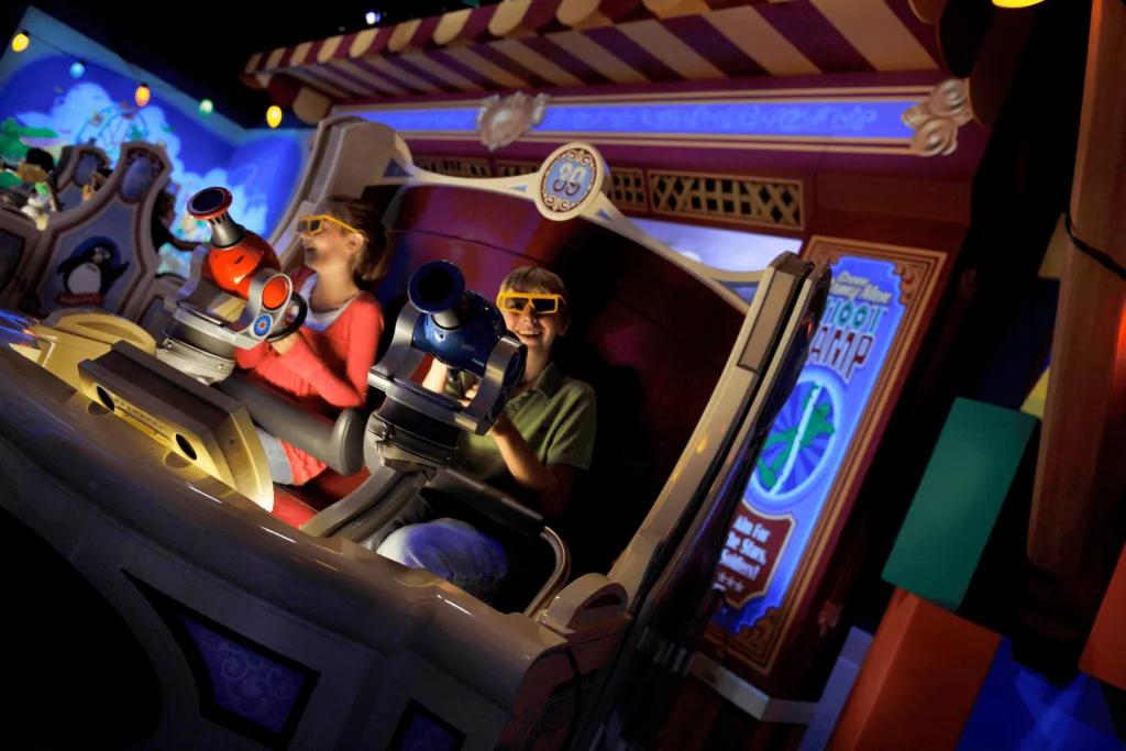 Kids sat in a circular ride cart wearing yellow 3d goggles and holding onto a red and blue shooter pointing them towards the screen showing toy story characters and targets.