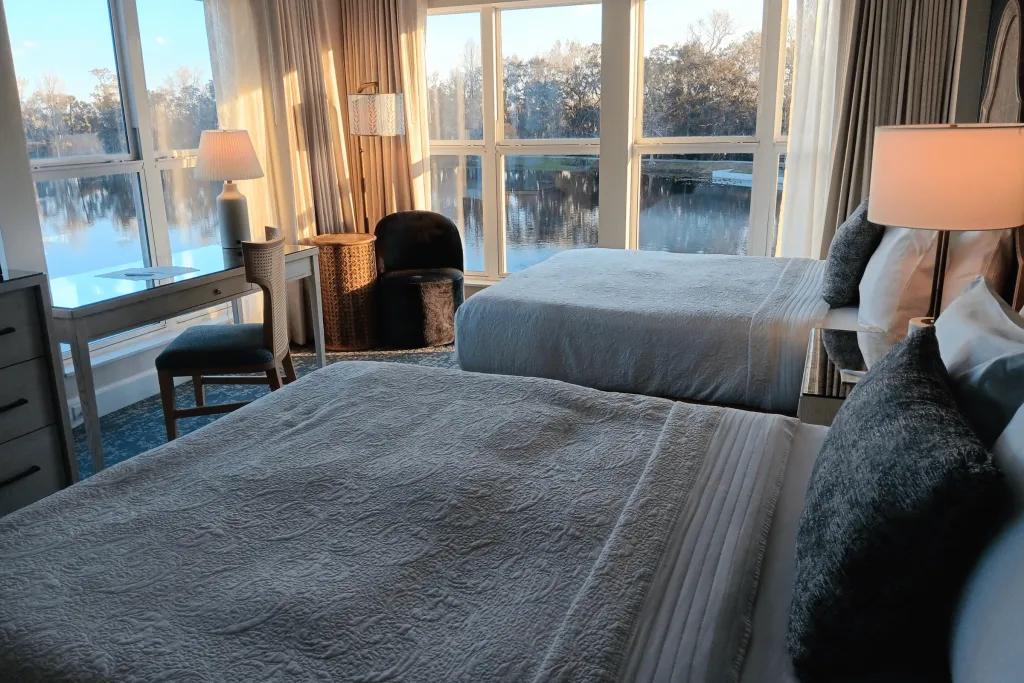 Two beds in a hotel room with patterned sheets and green pillows on facing out towards to 2 large windows from floor to ceiling overlooking the beautiful lake.