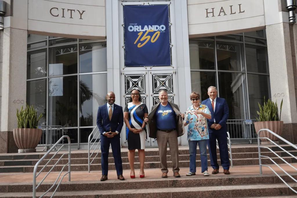 Mayor stood in a shirt celebrating the 150 year anniversary of ORLANDO with four other people. The City hall is behind decorated in banners saying Orlando 150.