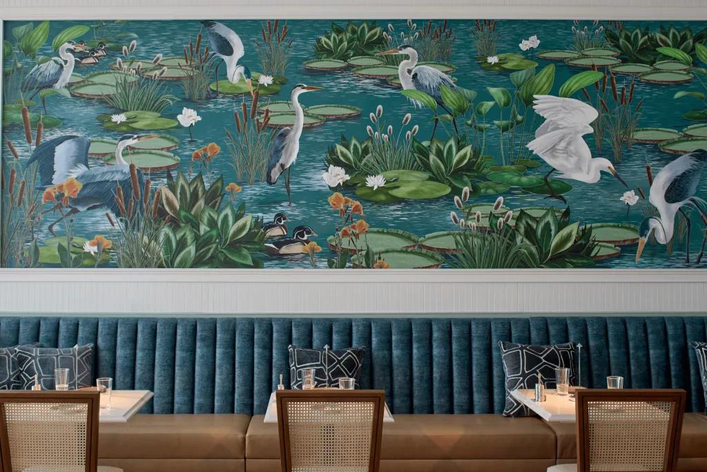 A restaurant with small table decorated with glasses, there is a booth side and a chair for each small table. There's a large wall behind full of art of herons in a lake.