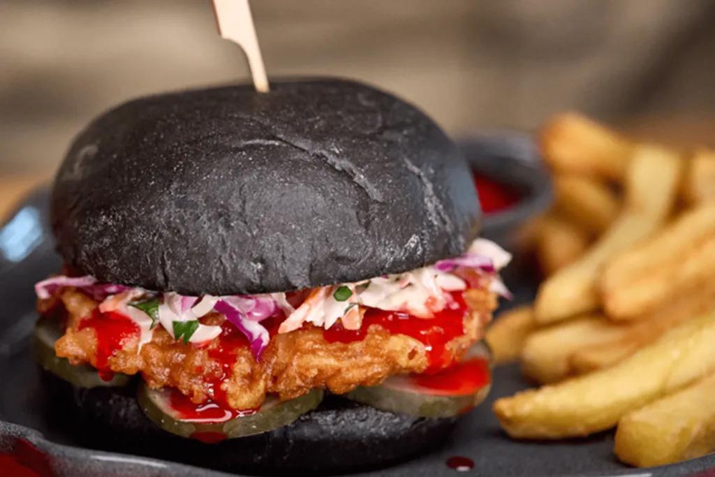 Black burger bun with fried chicken inside, coleslaw, onions, pickles and a bright red sauce. On the side there are fries and a red dip.