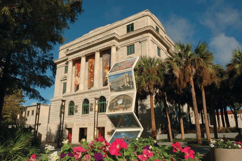 A large cream building with pillars and old architecture and a statue outside wit h historic images on and flower beds.