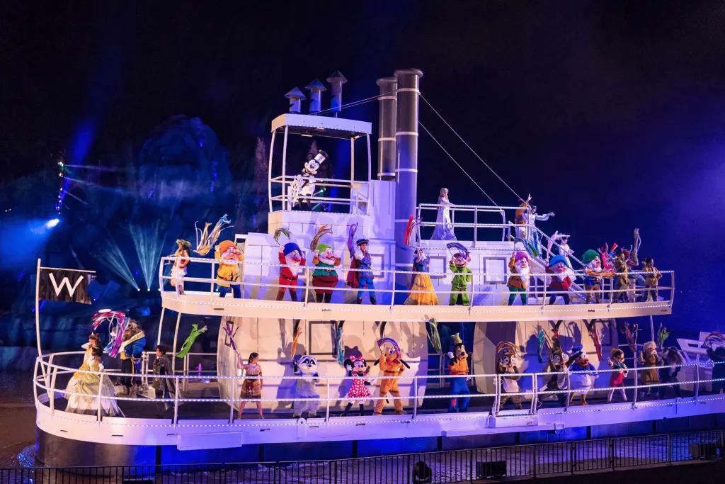 An old white steamboat full of almost all of the Disney characters, including all princesses, cartoon characters and Mickey driving the boat over a lake. They are all waving a colourful prop.