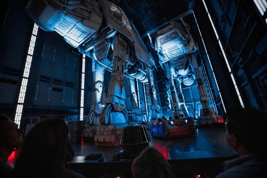 There are large robots with four legs and storm troopers sat inside. The rollercoaster carts are small with two rows of four and a robot sat in the front.