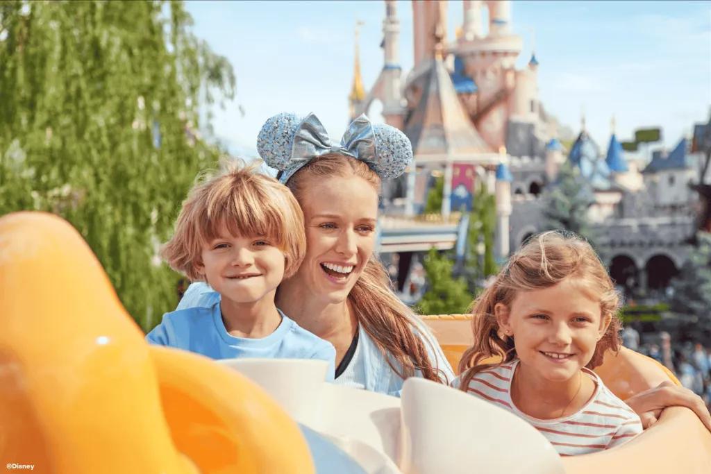 A woman and two young children in a ride vehicle shaped like Dumbo, in front of Sleeping Beauty Castle