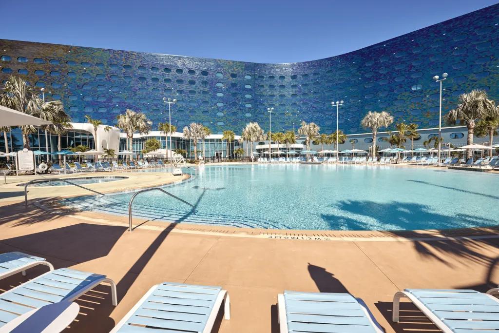 Large hotel with blue and purple ombre tiles on the outside, with a large pool outside surrounded by white deck chairs and palm trees.