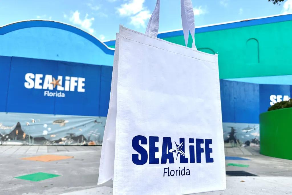 Huge blue building with SEA LIFE logo on it and a star fish. Under there is Florida label. There is a white tote bag being held up in front with a matching logo on it.