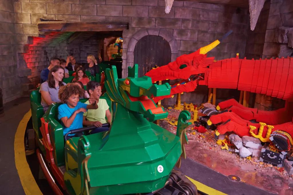 Kids and parents on rollercoaster, the car is a lego green dragon and it is going through a room with a large red dragon inside guarding jewels in a castle.