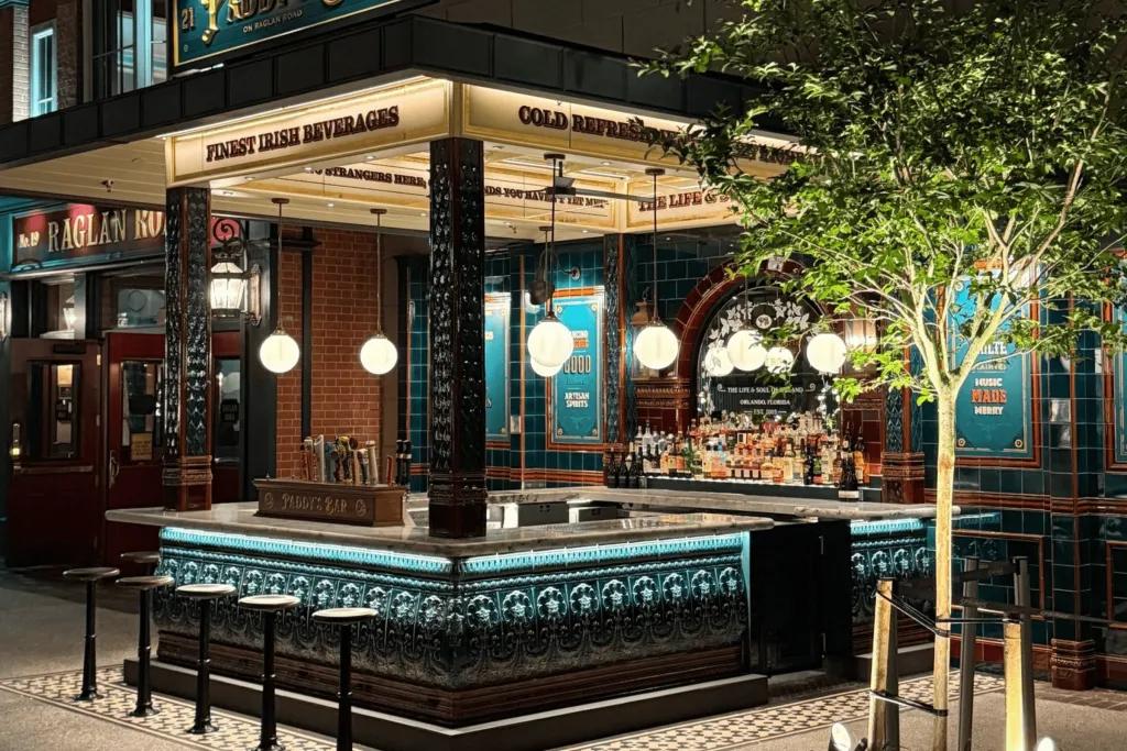 An outside bar with a modern, slick design. There are dark green/turquoise tiles on the wooden panelling and stool chairs to match. There are also low hanging circular lights below the sign.