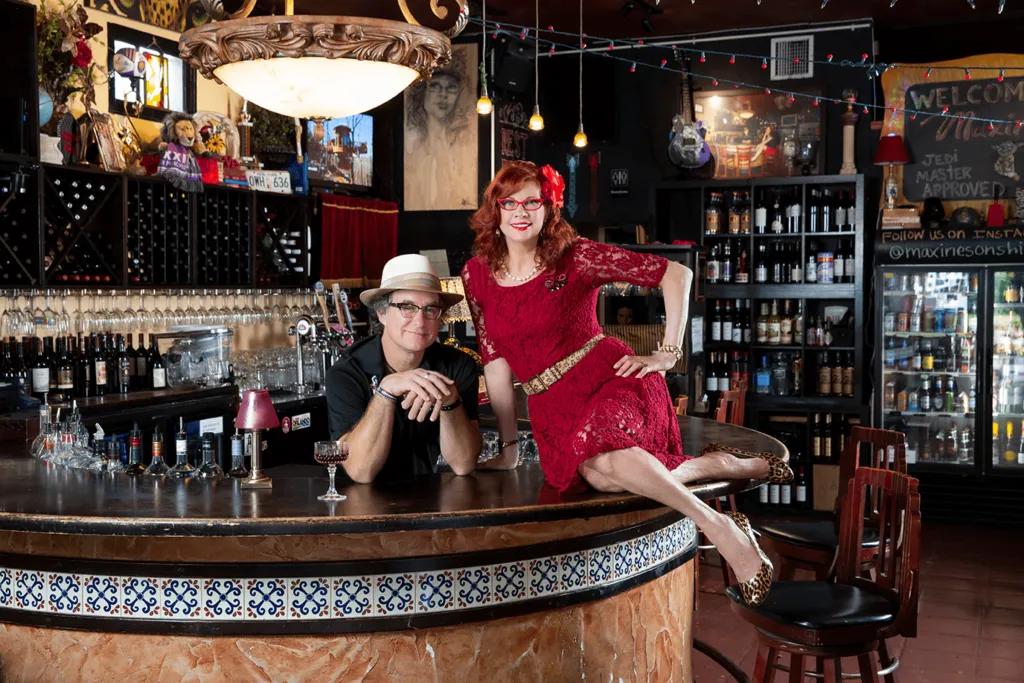 Woman in red dress with red flower in her hair sat on oval shaped bar, with bar stools behind her. A man is stood leaning on the bar from inside it. 