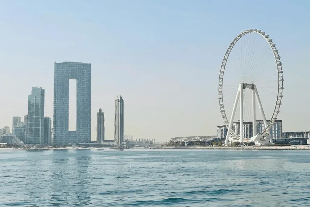 A large observation wheel on a beach in front of the sea and in the background is a frame shaped building and other unique architecture.