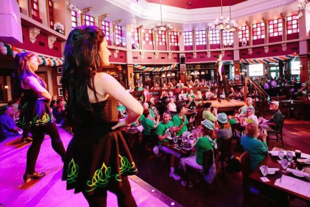 Two irish dancers on stage with round tables on the floor full of people dressed in green and Irish attire. 