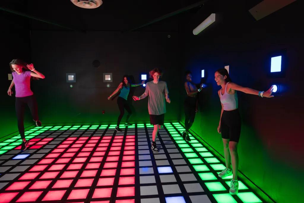 People in a room with tiles on floor lighting up different colours. People are stood on the green tiles and avoiding the red tiles.