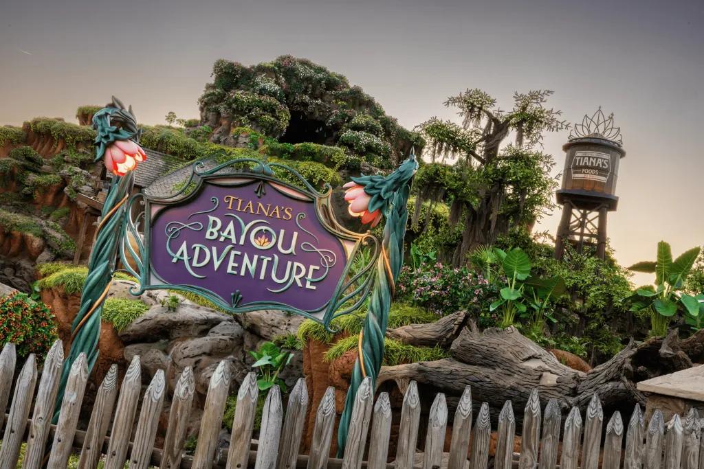 A sign made out of statue flowers which says "Tiana's Bayou Adventure". In the background there is a wooden water mill with a tiara on top. There is also a log flume ride covered in plants and flowers.