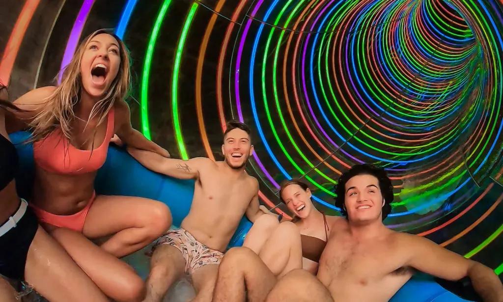 People sat in a large round rubber ring facing one another in a slide tube that is rainbow coloured with neon colours.