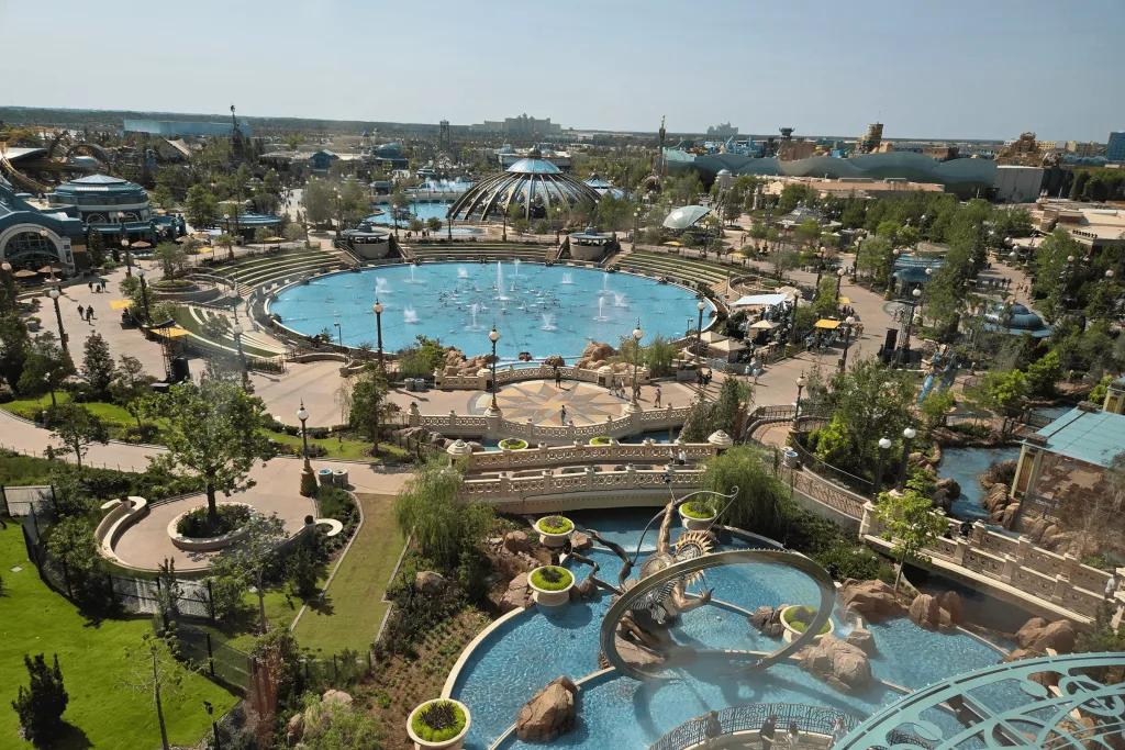 Large open space with two huge lakes, one featuring fountains, and pathways between the two. In the background you can see the Constellation Carousel.