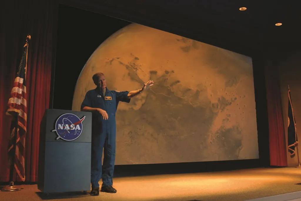 Man stood in space suit on stage with planet presented on screen behind him.