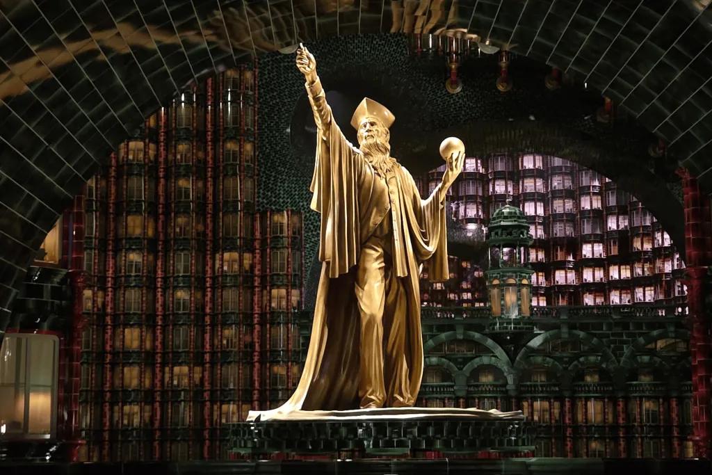 Golden statue of a wizard holding one hand up and the other is holding a golden ball. In the background is the Ministry of Magic.