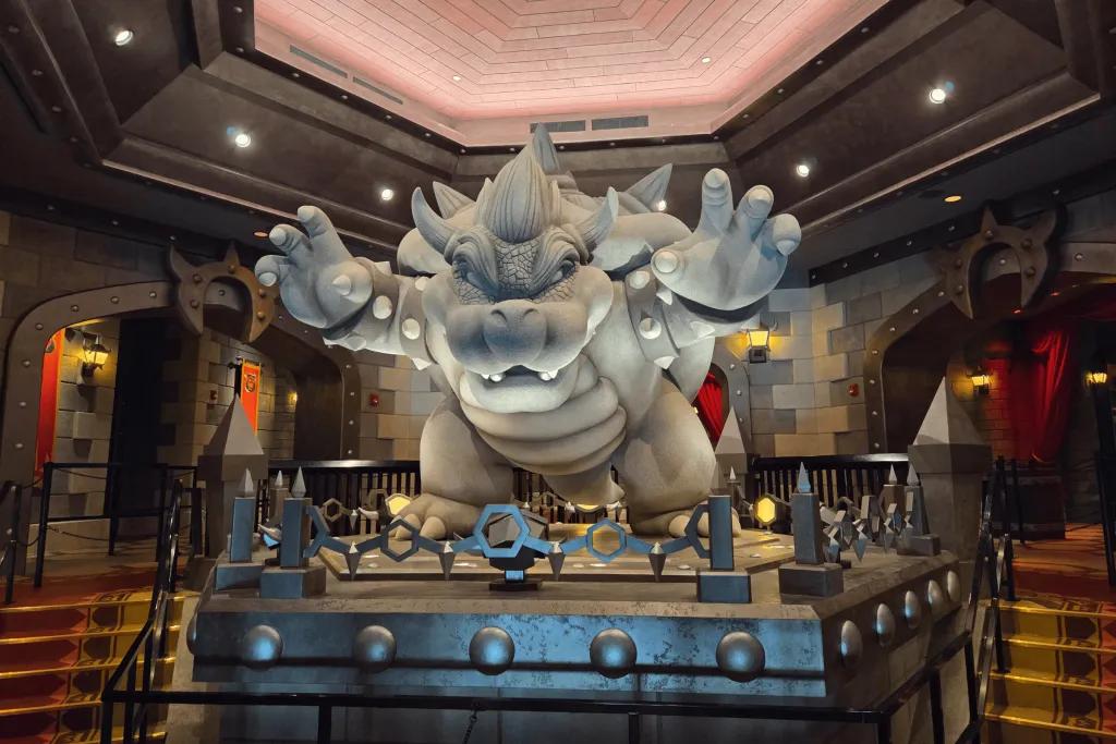 Statue of a weir creature called Bowser from Mario Kart, a turtle like creature with shell but the face is a bit scarier. The queue wraps around this statue.