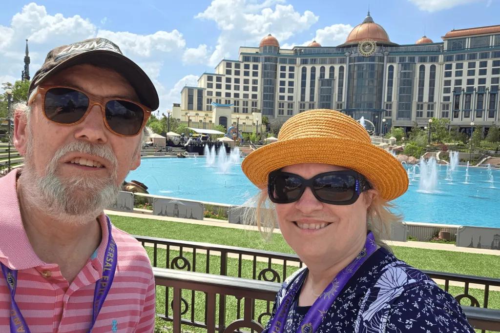 Man and woman dressed in summer outfits in front of Universal Helios Grand Hotel and fountains.