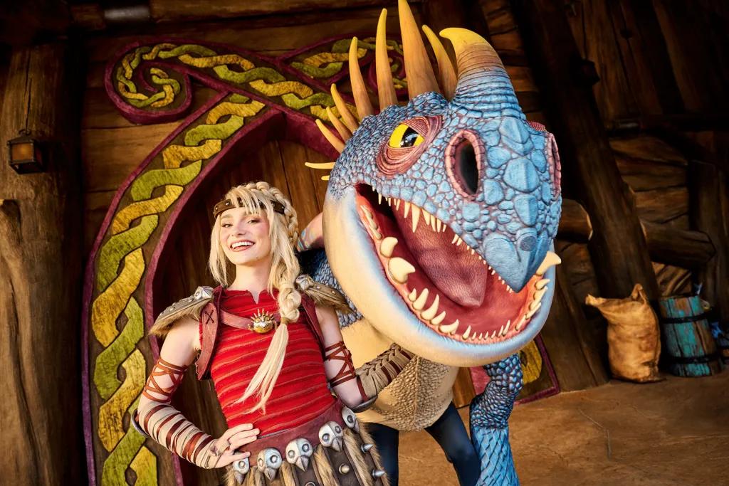Astrid, a blonde Viking girl wearing skirt with skulls on it stood next to a blue and yellow dragon.