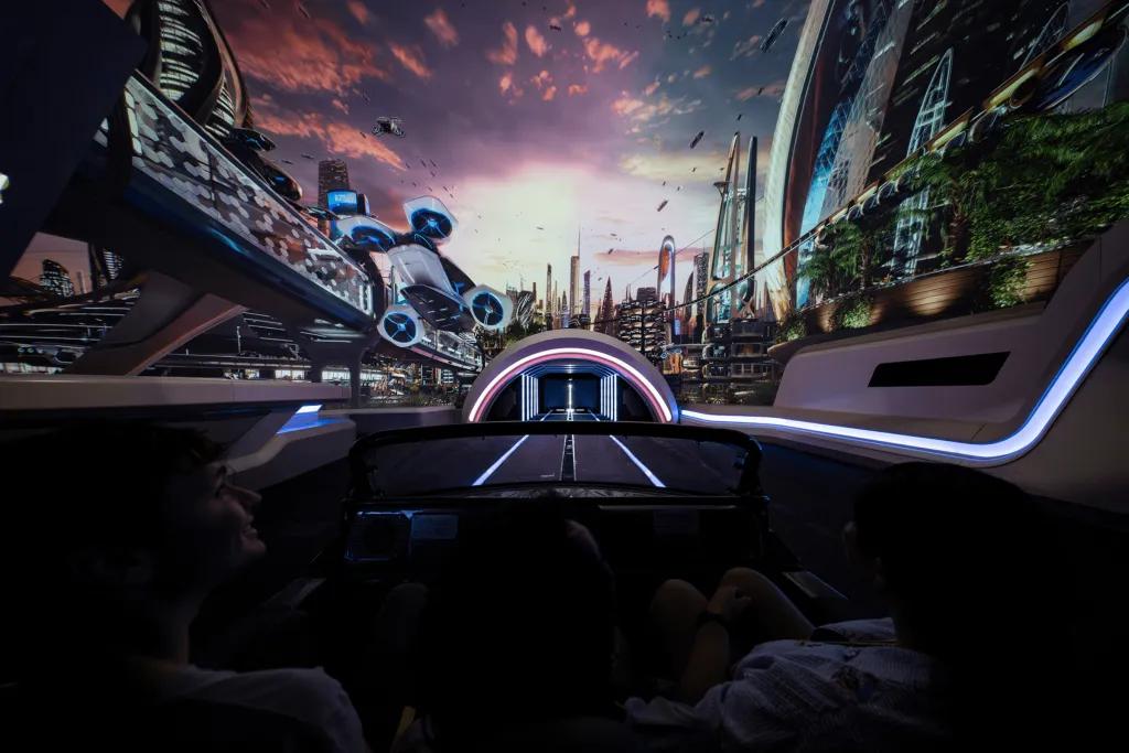 People sat in a car shaped vehicle, 4 in a row, there is a large screen with a modern, futuristic world on it and a small tunnel where the track leads to.