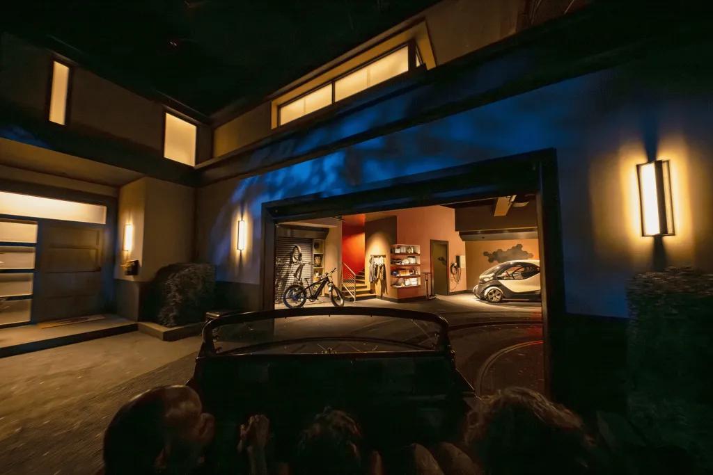 People sat in car shaped rollercoaster vehicle heading into a room where there are bikes and other things stored.