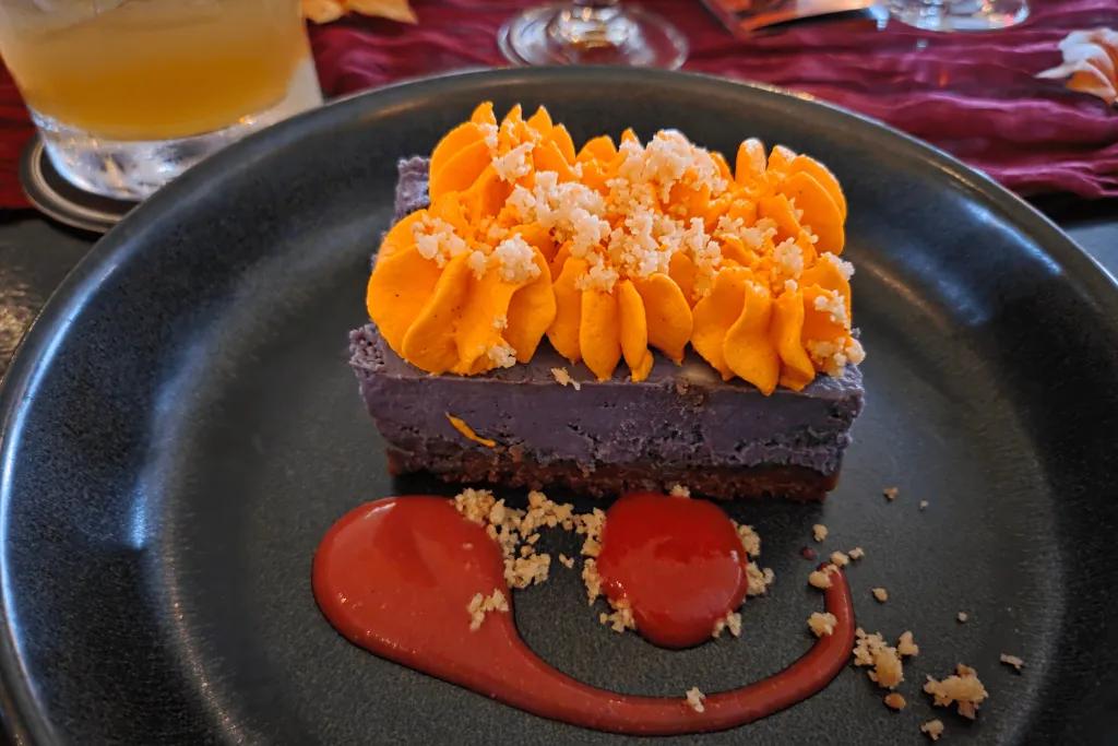 Purple cheesecake with orange icing and a red sauce with some biscuit crumbs on top.