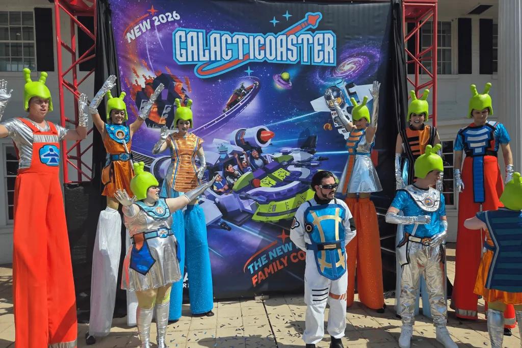 People with alien green helmets on and alien outfits, some stood on stilts, stood in front of a ride poster.