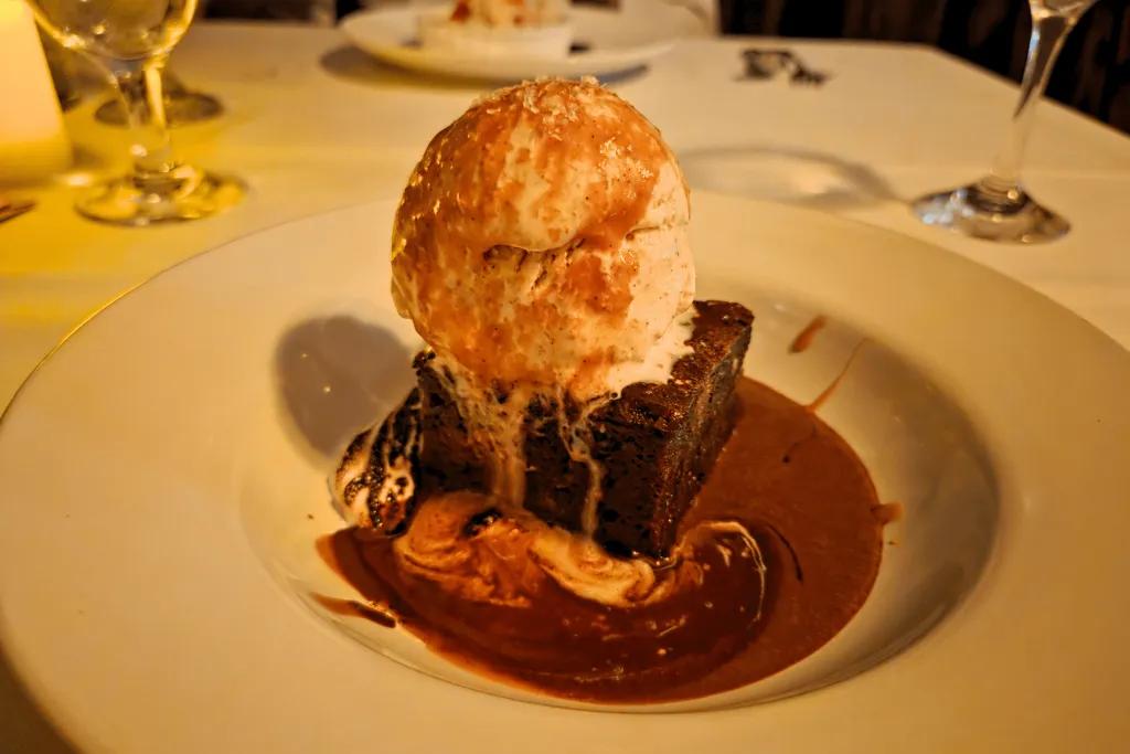 Large square brownie in a chocolate sauce with a large scoop of ice cream on top and caramel drizzled over.