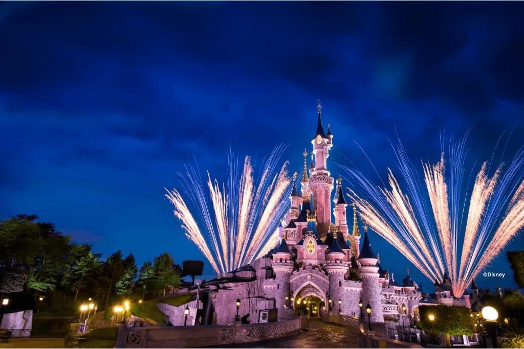 Gemini said Illuminated pink castle at Disneyland Paris with fireworks against a deep blue night sky.