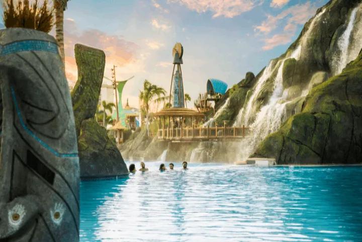 People relaxing in a large blue swimming pool at a tropical-themed water park, featuring a giant artificial volcano with waterfalls and carved tiki structures in the foreground under a sunset sky