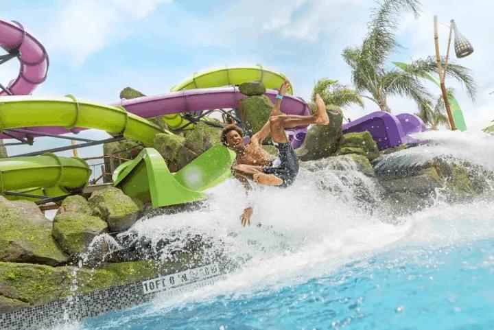A person splashing mid-air as they fly out of a green water slide into a deep pool, with twisting purple and lime green slides and artificial rocks in the background
