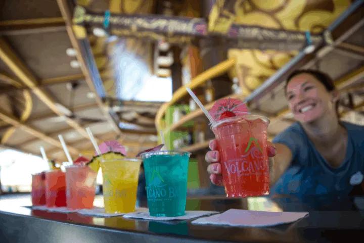 A smiling bartender handing over a pink tropical drink with a small umbrella, with a line of colorful cocktails in plastic cups—including blue, yellow, and red—sitting on the bar counter