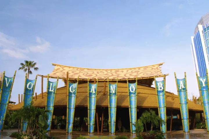 The entrance of Volcano Bay water park showing a large thatched-roof building with tall blue banners that spell out "VOLCANO BAY" under a bright blue sky