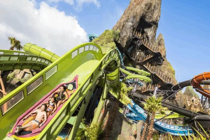 A family of three riding a multi-person raft down a bright green water coaster track, with a massive artificial volcano and various tangled water slides in the background under a blue sky