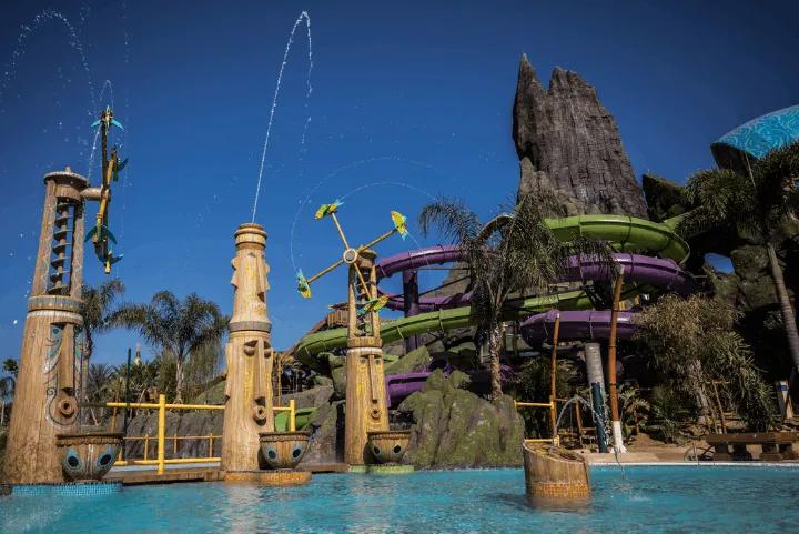 A water park splash pool featuring carved wooden tiki poles spraying water streams into the air, with twisting purple and green water slides and a large artificial rock volcano rising in the background against a clear blue sky