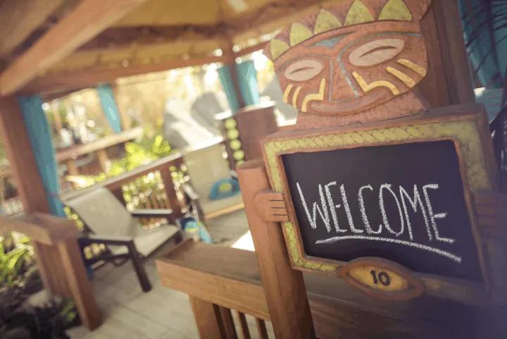 A close-up view of a wooden tiki-themed welcome sign with a chalkboard reading "WELCOME" and the number 10, located at the entrance of a private outdoor cabana with lounge chairs inside