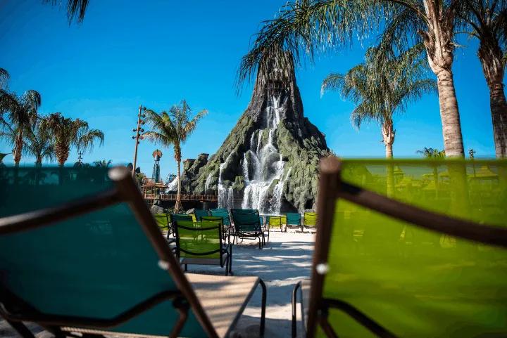 A view of a large artificial volcano with streaming waterfalls, framed in the foreground by empty teal and lime green beach lounge chairs on white sand under palm trees and a clear blue sky