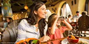 A mum and young girl sat at a table eating dinner. The girl is using binoculars to look out the window