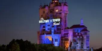 A large creepy looking tower standing tall above trees which looks like a run down, creepy hotel with a sign lit saying the Hollywood Tower Hotel.