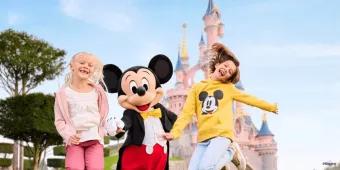 Zwei junge Mädchen springen mit Micky vor dem Schloss von Disneyland Paris