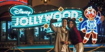 A couple taking a selfie in front of a neon sign for Disney Jollywood Nights