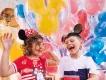 A young girl wearing a headband with Minnie Mouse ears and a young boy wearing a hat with Mickey Mouse ears standing in front of multi-coloured balloons