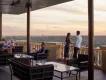 Terrace with brown chairs and glass tables on, there are two groups of two people sat to the left of the image and a couple stood up overlooking the view of the terrace.