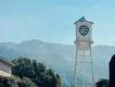 White water tower with blue shield shaped logo with letters WB on. In the background there is a large tree and big mountains with a blue sky overhead.