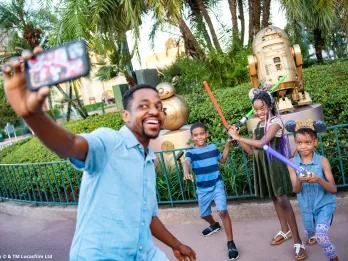 A father and three children taking a selfie posing with lightsabers in front of the Star Wars golden statues